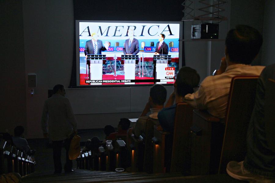 In a dim lecture hall, students watch a live video feed of three people at podiums captioned “REPUBLICAN PRESIDENTIAL DEBATE.”