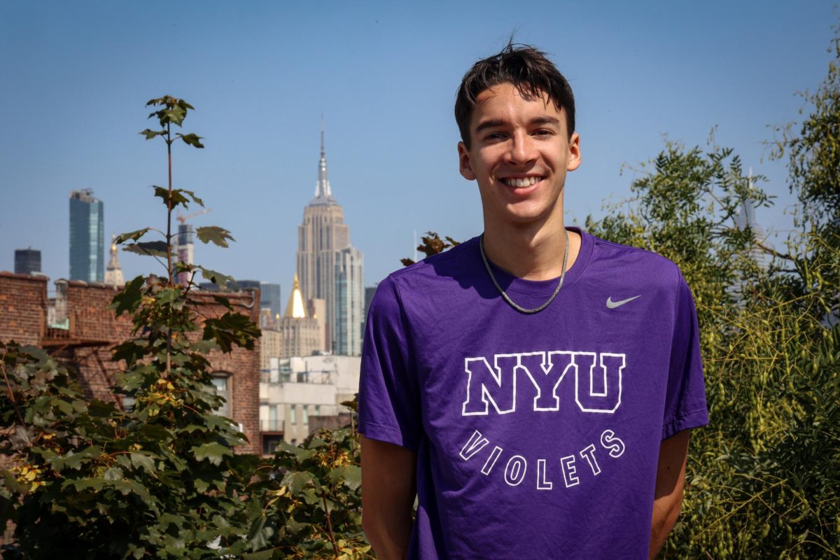 Athlete Julian Aske wears a purple short sleeve shirt that reads “N.Y.U. VIOLETS” in block white lettering as he poses on a roof with the Empire State Building in the background.