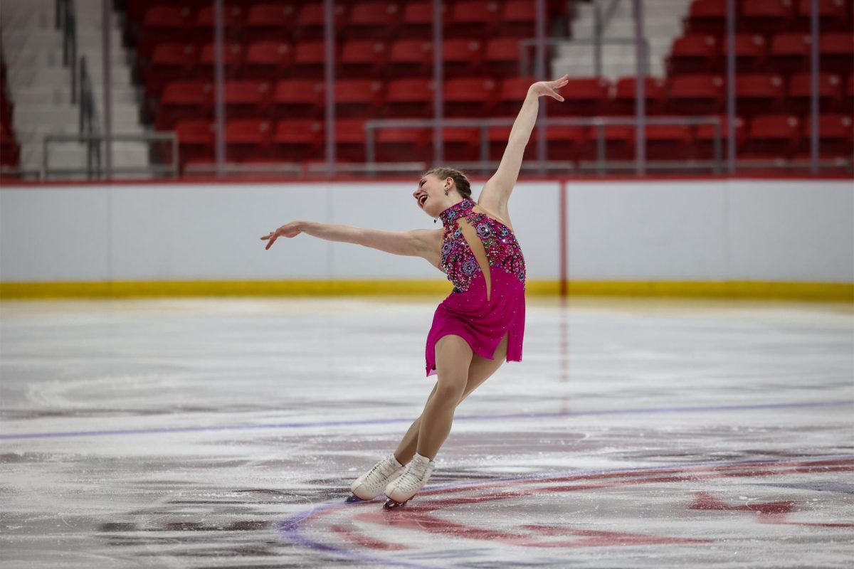 An ice skater in a pink leotard skates on the ice with her arms raised in a pose.