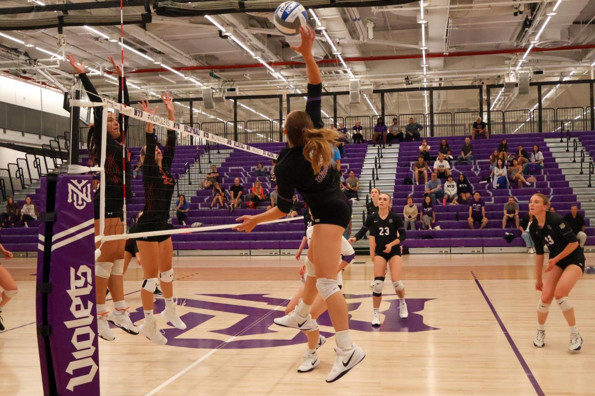 One volleyball player jumps up to spike the ball while her opponents on the other side of the net jump to block her.