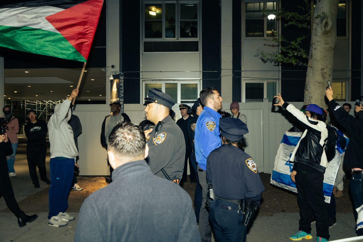 A group of police officers and protesters gathered outside a white building. One protester holds a large Palestinian flag on the left, while another has an Israeli flag on the right and police officers stand in the middle.