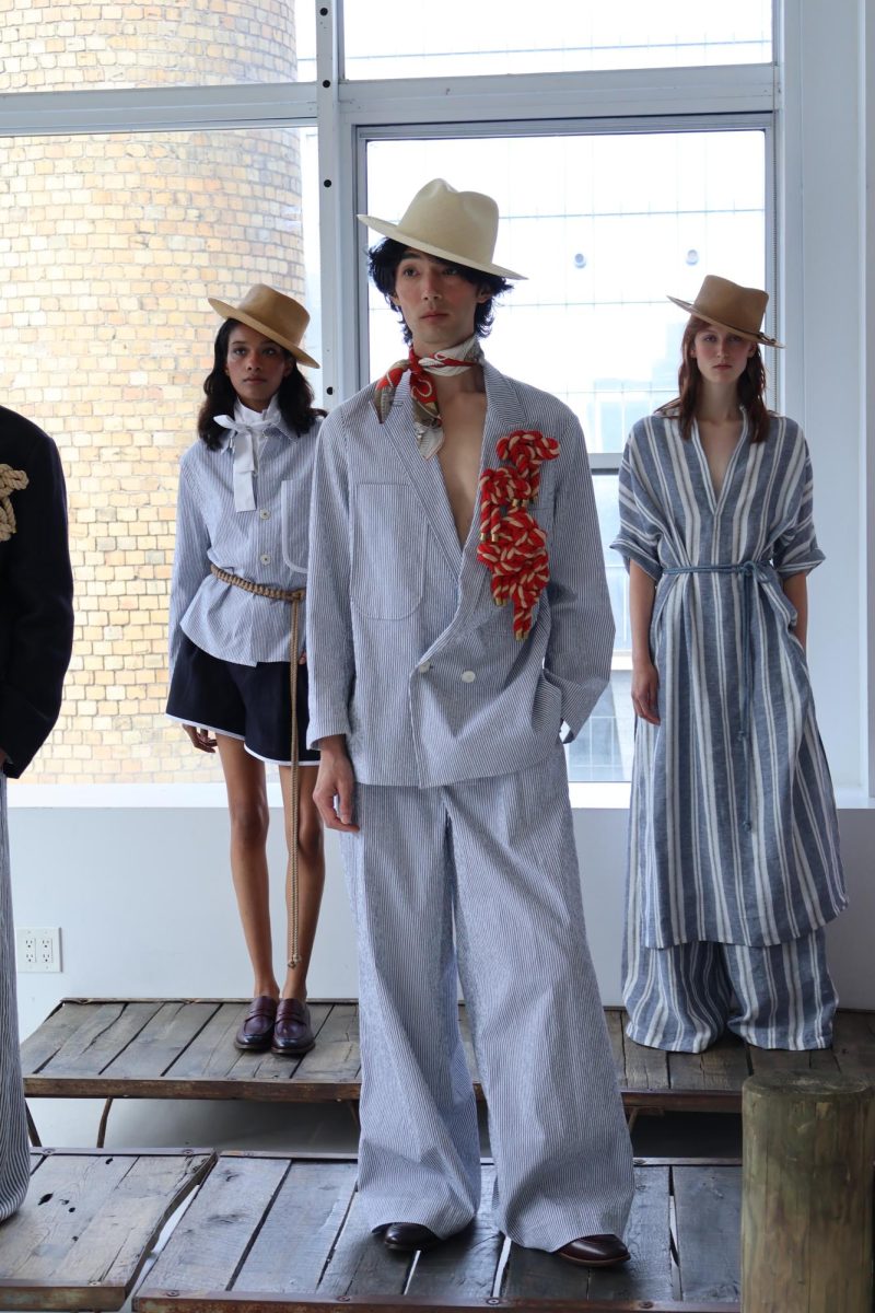 A model wears an oversized blue pinstripe suit and a beige hat. There is a red bandana tied around their neck and collection of small red knotted ropes attached to one breast of their jacket.