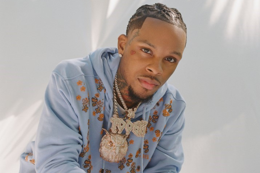 A man wearing a light blue hoodie and lots of gold jewelry sits in front of a white background.