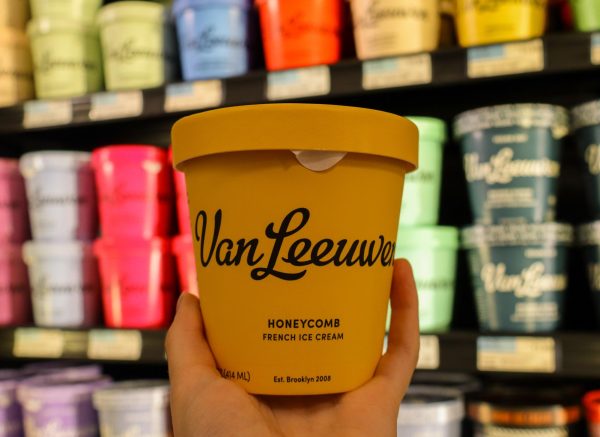 An orange tub of ice cream that reads, “Van Leeuwen Honeycomb Ice Cream” in black cursive lettering with other flavors on display in the background.