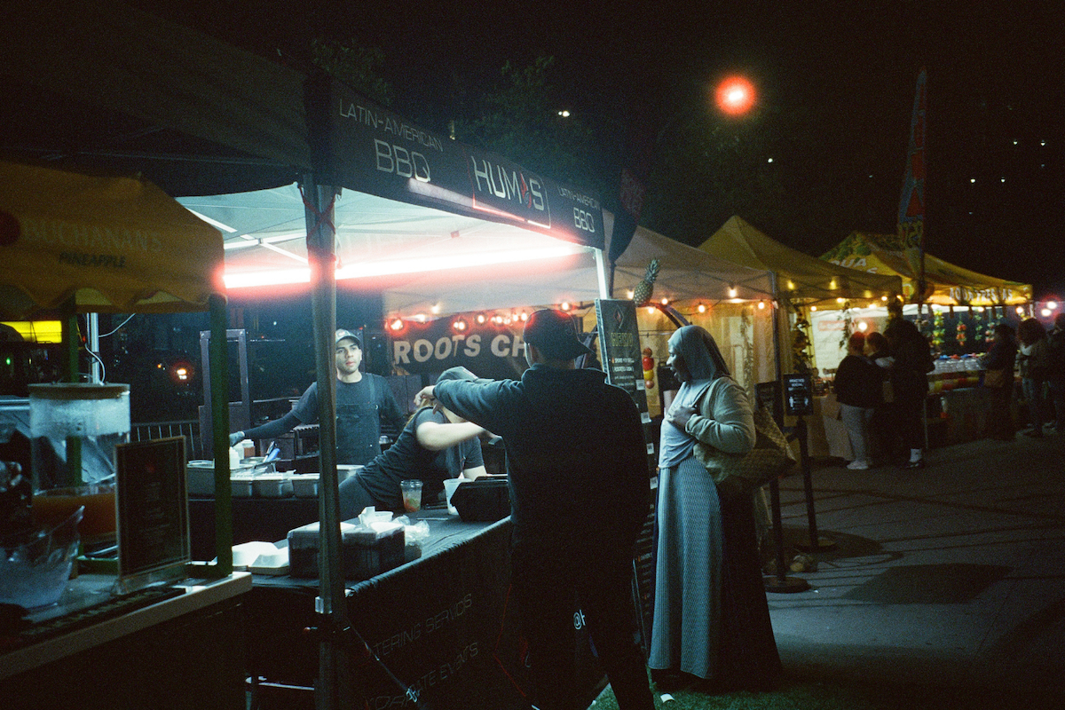 Someone ordering food at a tent with “LATIN-AMERICAN BBQ” and “HUMS” written on it.