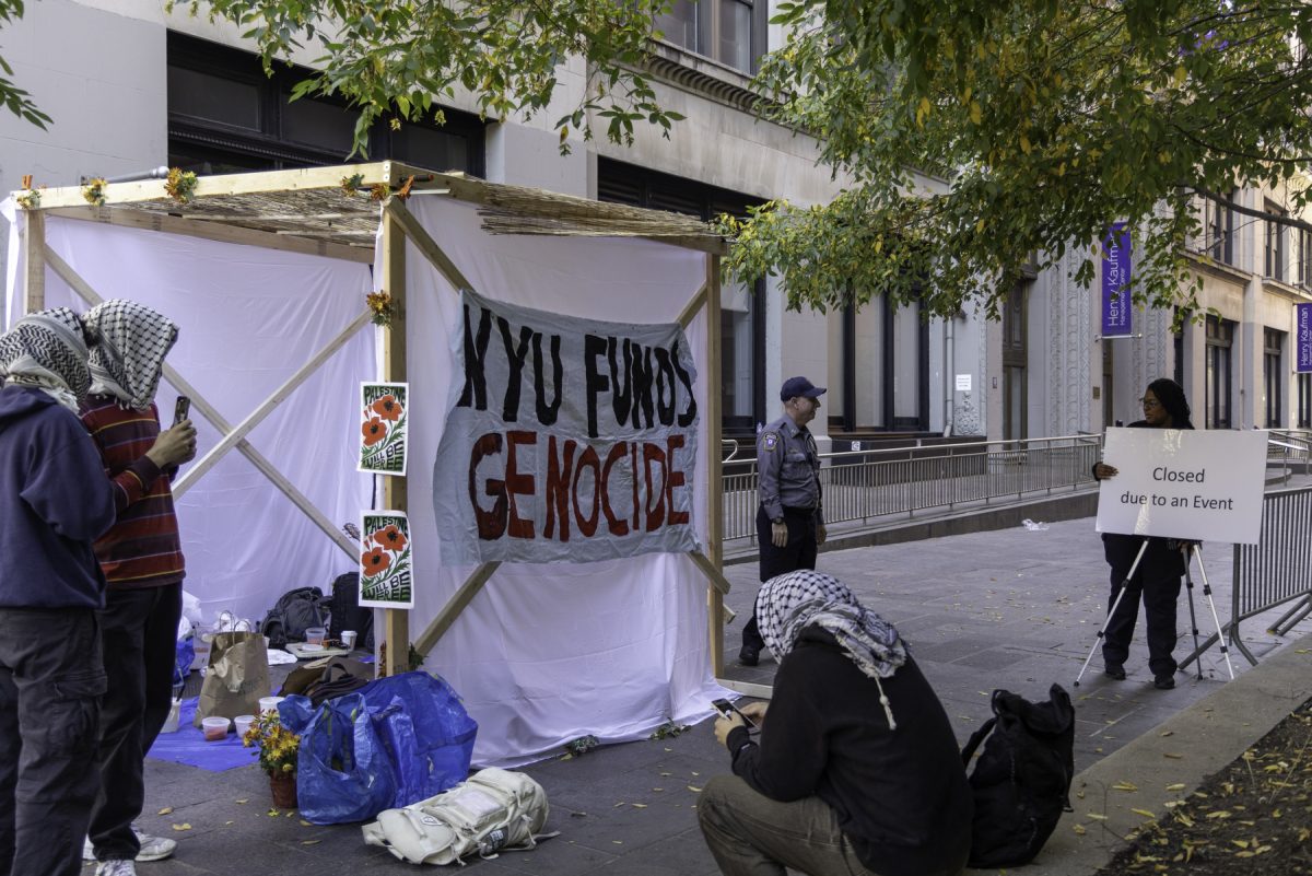 A structure with draped white sheets that has the words “N.Y.U. FUNDS GENOCIDE” painted in black and red paint sits in the middle of a walkway with students confronting university staff.