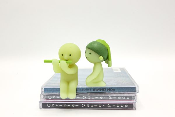 Two miniature green figures sitting atop a pile of CDs. The one on the left is playing the flute while the one on the right watches.