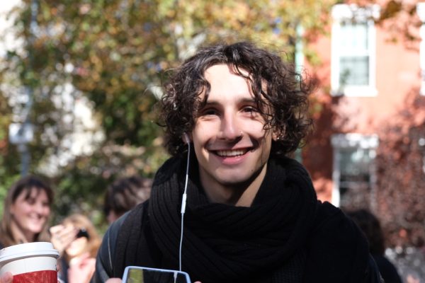 A close-up shot of a curly-haired brunette young man wearing a black scarf and black jacket, smiling at the camera. 