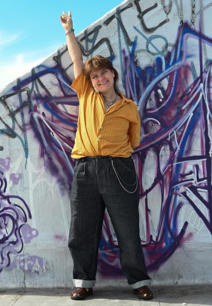 A person is standing in front of a graffiti wall, raising their hand in a V-sign. They are wearing a striped orange shirt and black chained pants.