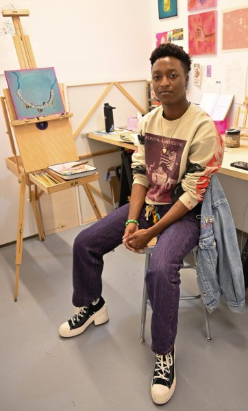 A person sitting in an art room filled with wood easels and paintings on the wall. They are wearing a yellow graphic sweater that says “REBELLION HOMOSEXUAL” and purple pants.