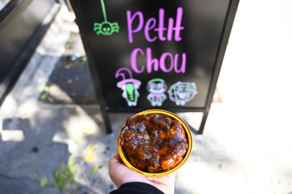 A round tin of a glazed dessert in front of a black sign with the words “Petit Chou” in lavender text surrounded by illustrations of cute halloween creatures.