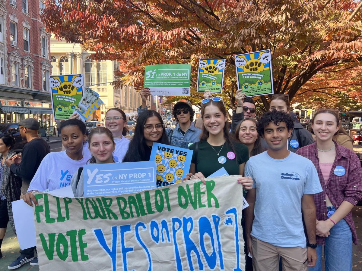 A group of people stand on the sidewalk. They hold flyers, posters and a large sign stating “FLIP YOUR BALLOT OVER VOTE YES ON PROP #1.”