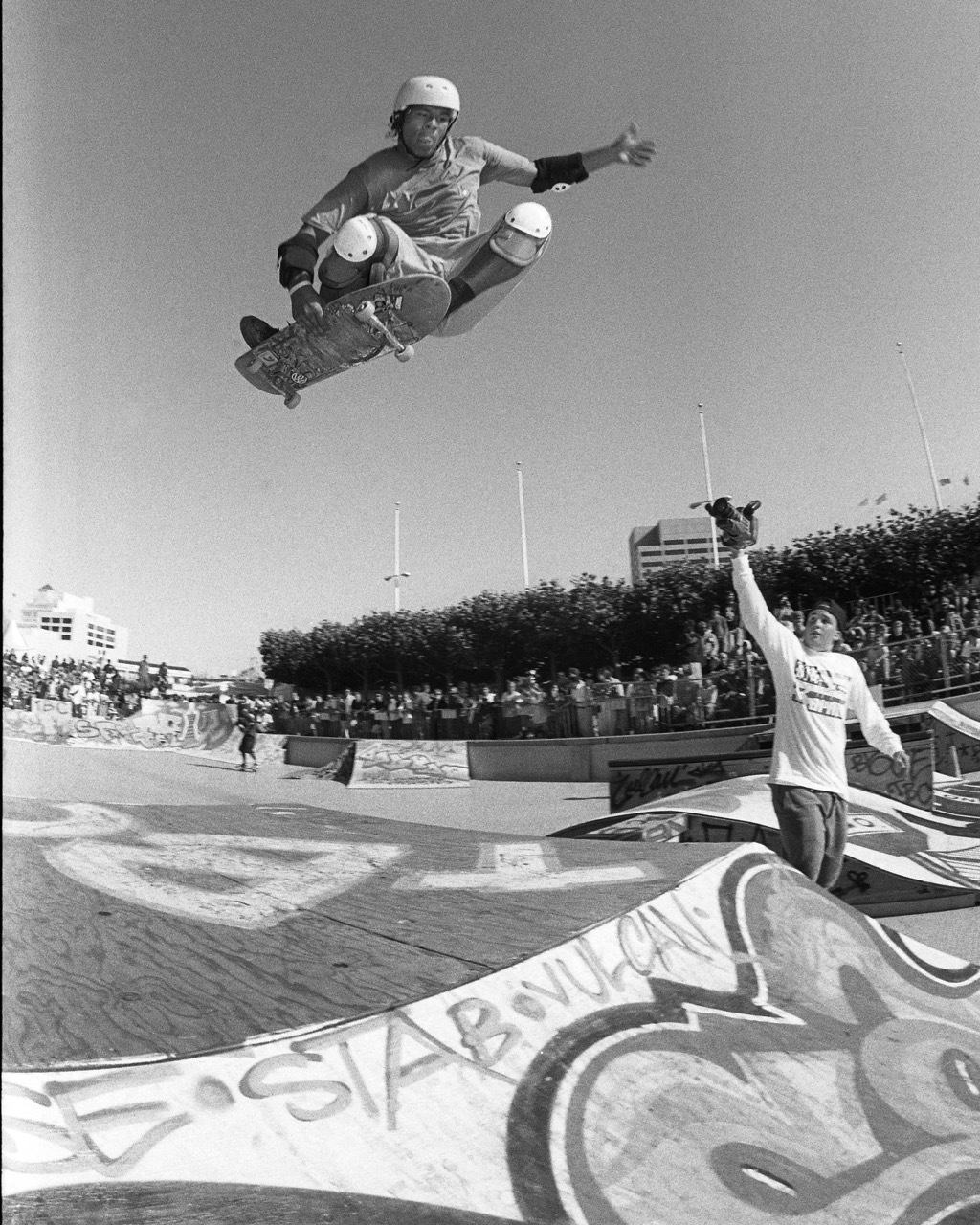 A black-and-white photo of a man jumping over the camera on a skateboard. There is a man behind him who is filming him.