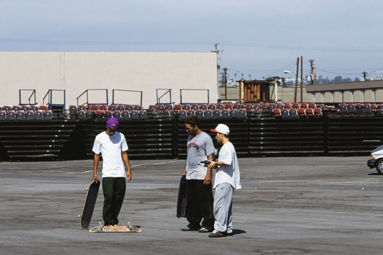 Three men standing in an empty parking lot. One man is wearing a white shirt, black pants and is holding a skateboard. Another man is wearing a gray shirt with black pants, and the other wears a white shirt with gray pants.