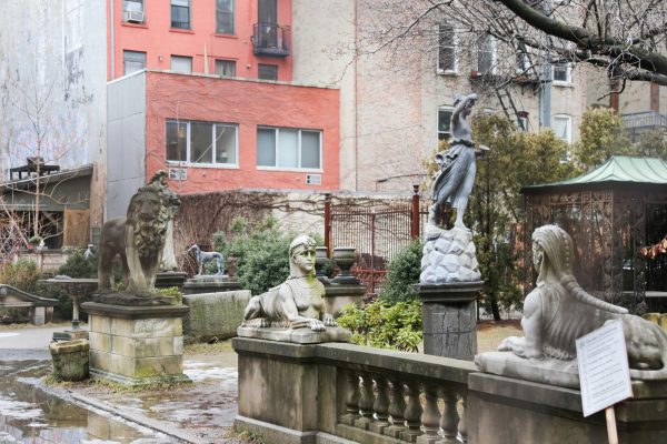 An area with a lot of garden statues including lions, sphinx, vasts and a female figure with urban buildings in the background. 