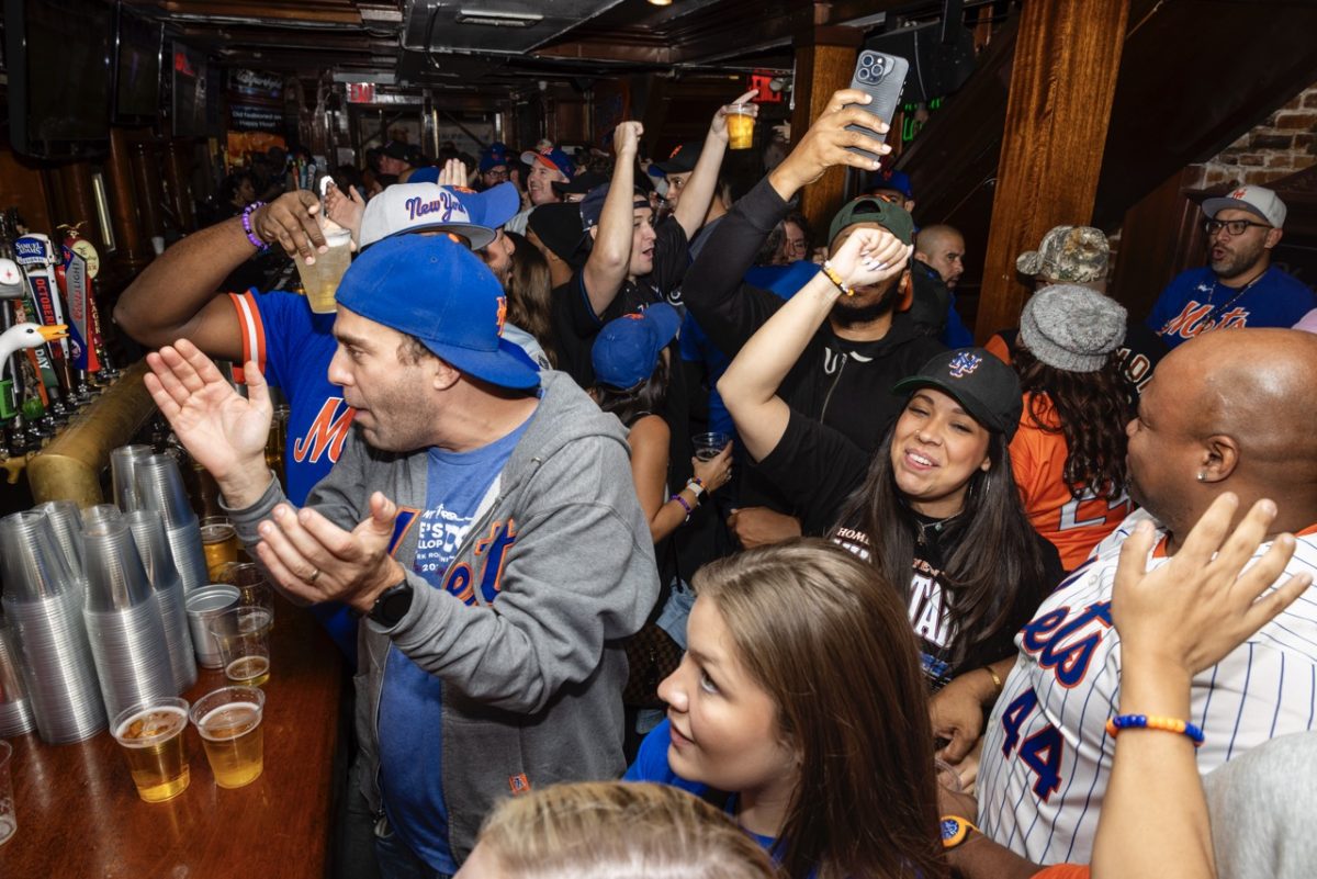 People in a crowded bar wearing Mets gear cheer and clap.