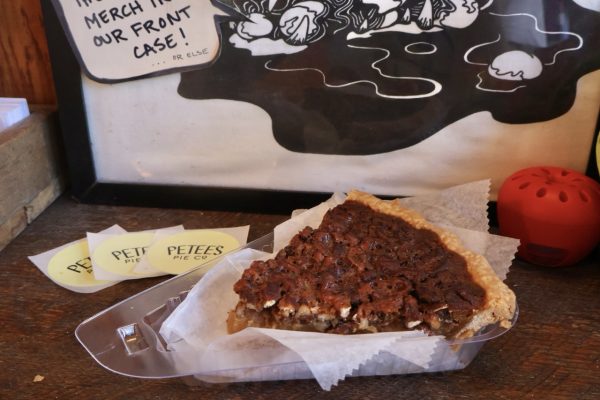 A slice of pecan pie in a plastic container on a wooden counter, with stickers from “Petee's Pie Co.” next to it.