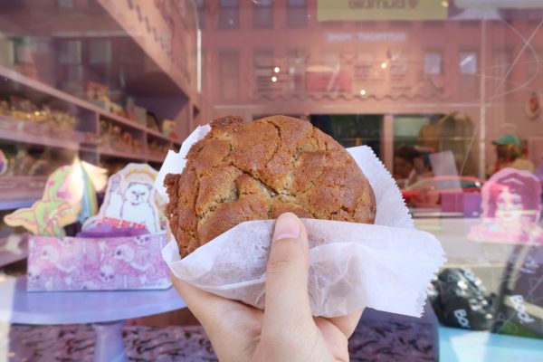 A hand holding a large brown cookie wrapped in paper in front of a pink bakery display window. 