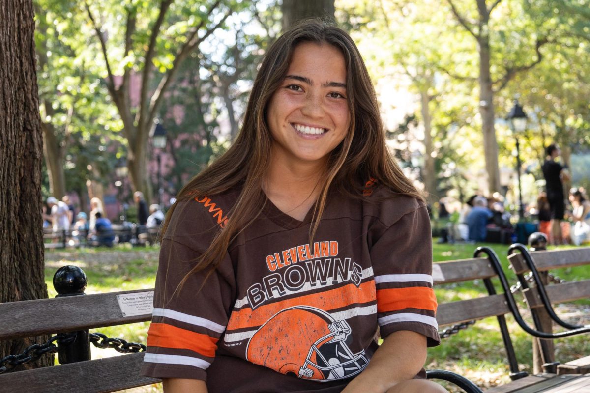 A girl with brown hair sits and poses on a bench in a park. She is wearing a brown shirt that says “CLEVELAND BROWNS”.