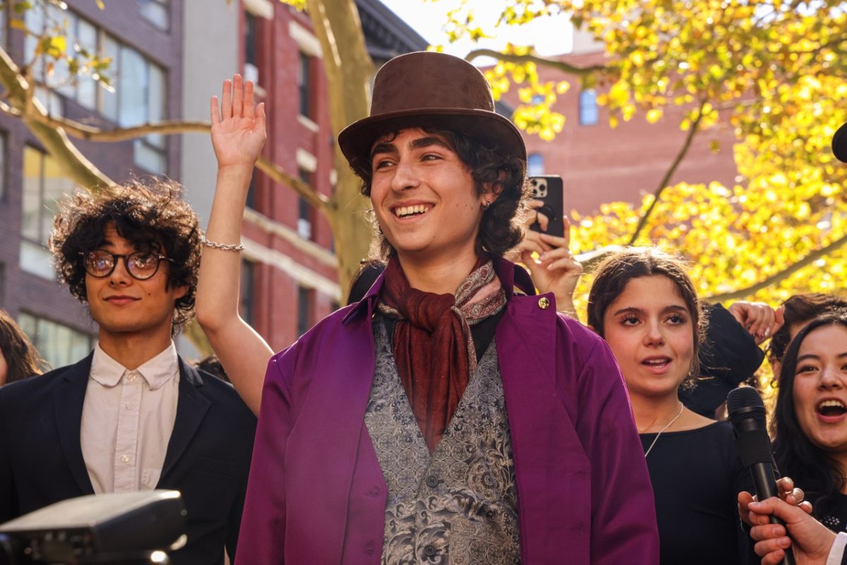 A look-a-like in a Willy Wonka costume smiling with a microphone in front of him while people behind him smile and raise their hands in the air.