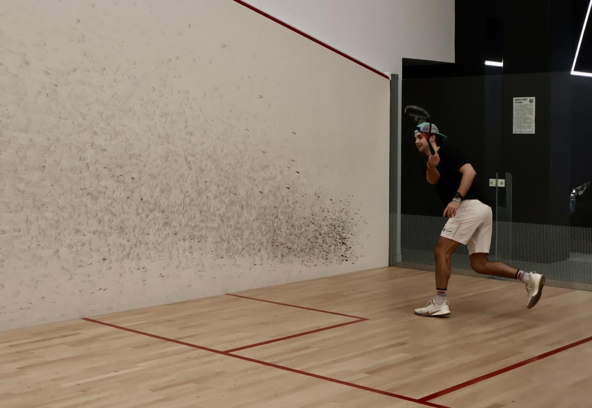 A man swings a racquet. Behind him is a wall with black marks from the ball.