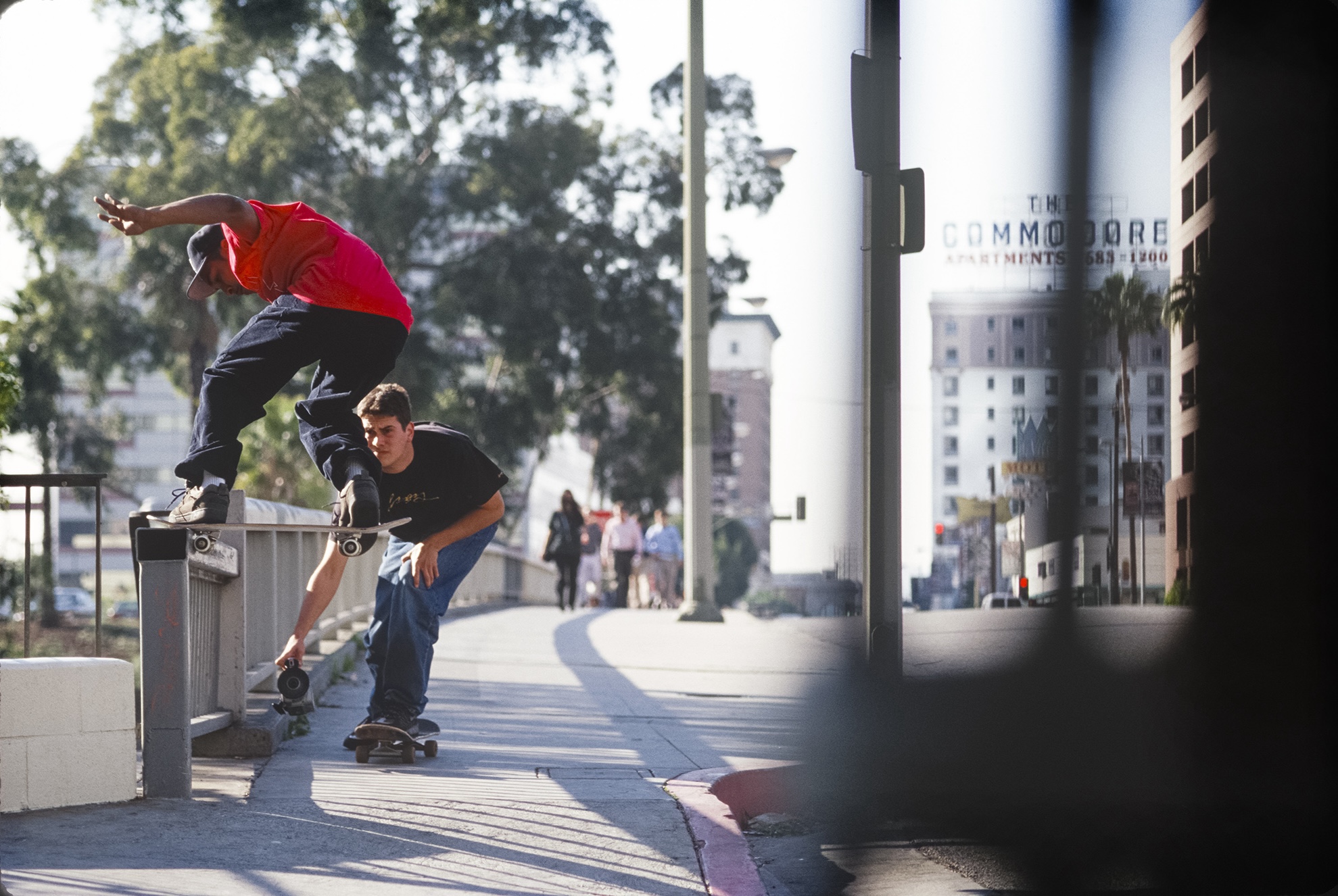 A man in a red shirt and black pants jumping on his skateboard on a fence. A man skateboards behind him and films him with a camera.