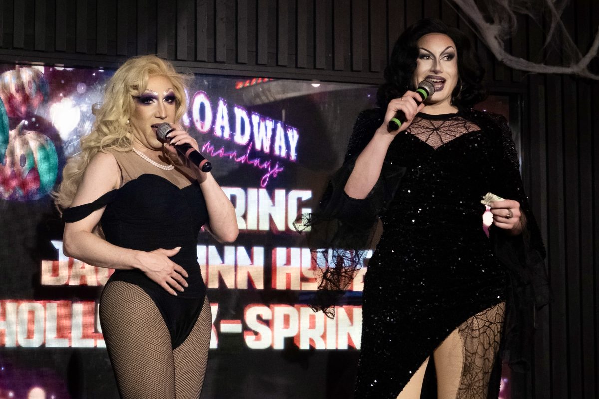 Two drag queens stand on stage holding microphones. One wears a blonde wig and a black leotard, and the other wears a black wig and a long sparkly black dress.