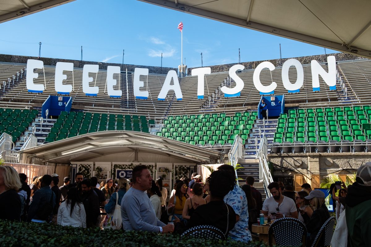 A stadium is packed with people around tables and chairs. In the stands, above the scene, are large white and blue letters spelling out “EEEEEATSCON.”