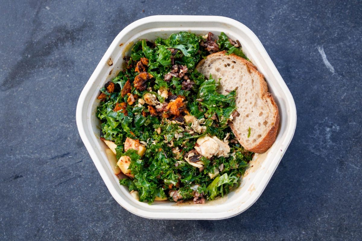 A top-down view of a hexagonal bowl containing dark green vegetables, meat and other ingredients tossed together with a slice of bread on top.