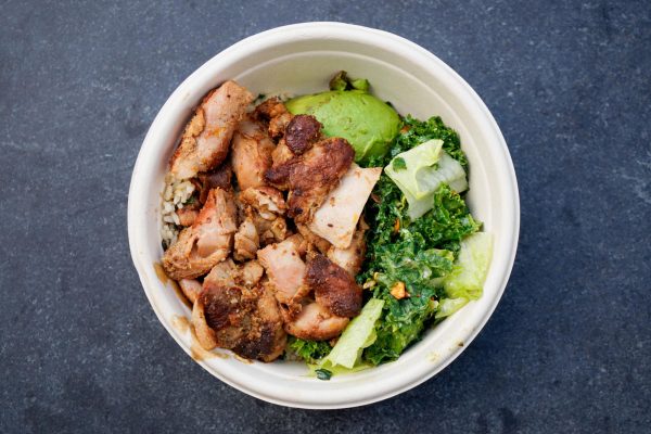 A top-down view of a bowl with chicken, dark green vegetables, avocado and other ingredients on a dark gray surface.