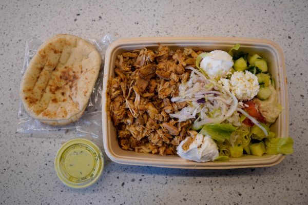 A top-down view of a rectangular bowl holding chiken, vegetables and other toppings, with a flat bread and container of dressing on the side.