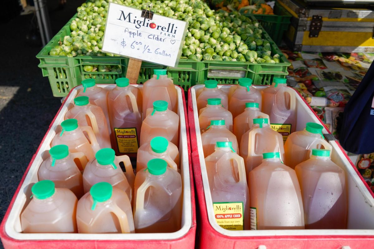 Crates of bottled apple cider labeled “Migliorelli Farm” and “$6.00 ½ Gallon” in front of a crate of brussel sprouts.