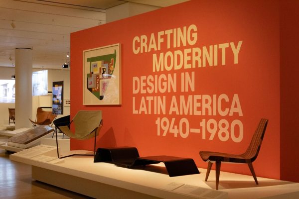 An exhibit featuring modern furniture on a stage with a large red panel that says “CRAFTING MODERNITY DESIGN IN LATIN AMERICA 1940-1980.”