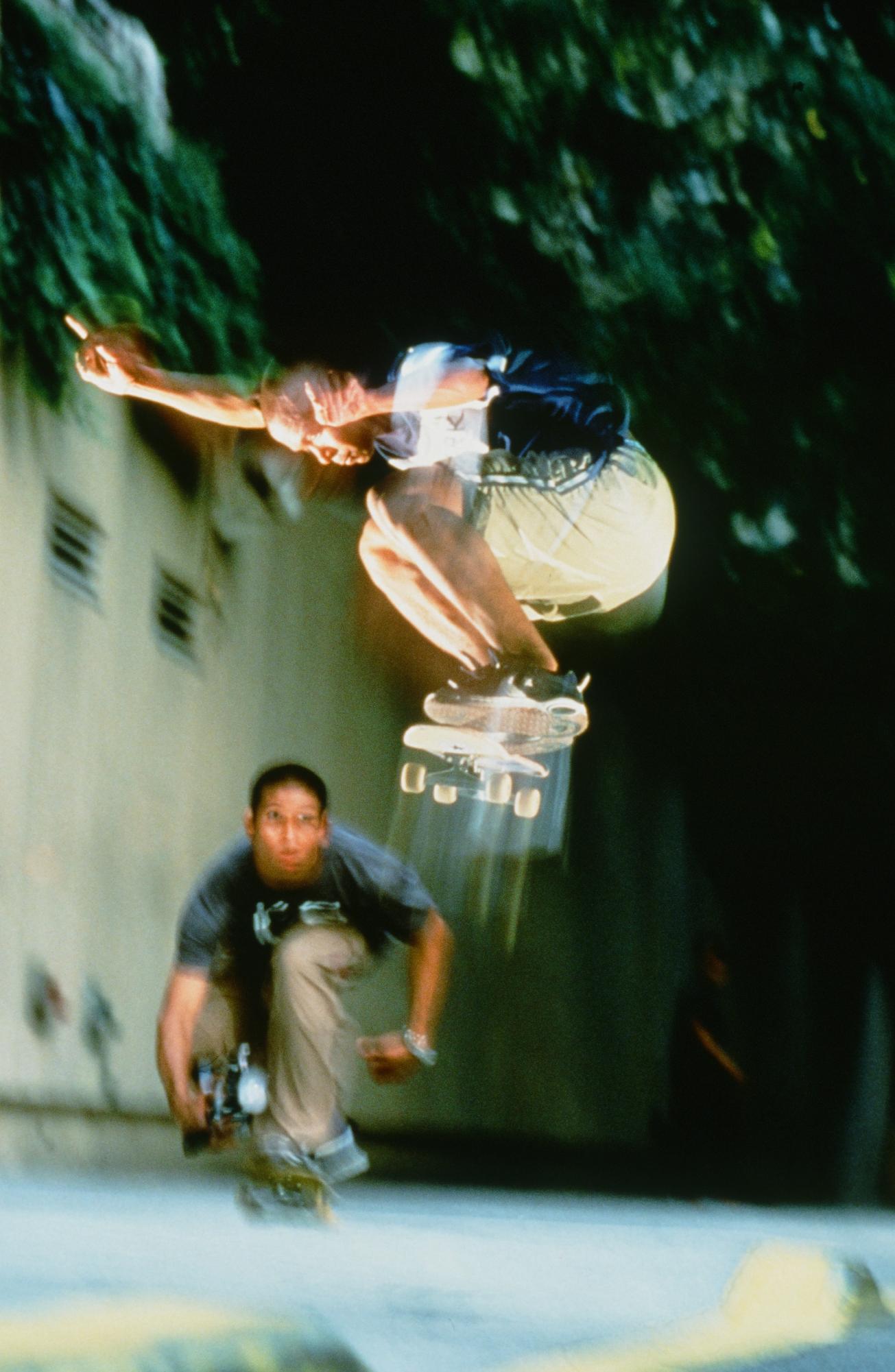 A blurry image of a man jumping on a skateboard. There is a second man squatting on a skateboard behind him.