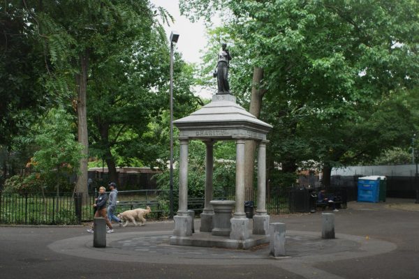 A green park surrounded by trees with people walking around. In the middle is a white monument that contains a statue on top.