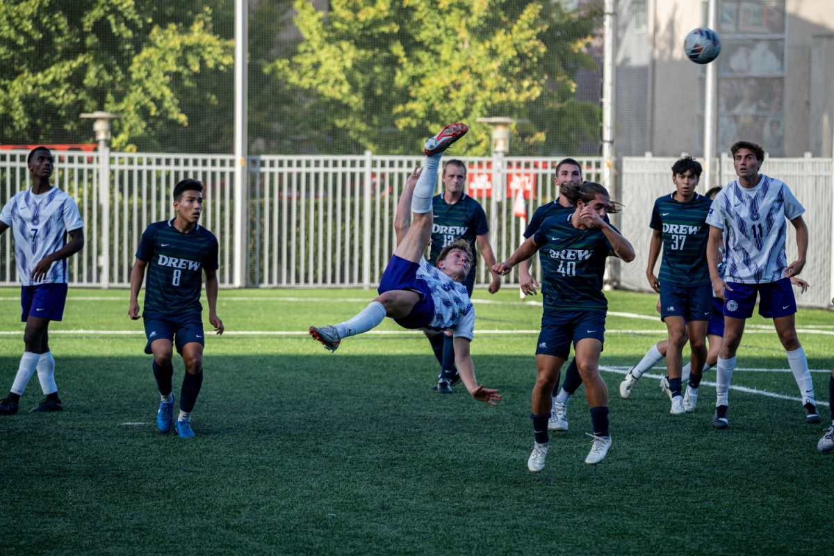 A soccer player in an N.Y.U. jersey is mid-air, with his leg above his head as he is about to kick a ball. He is surrounded by players of the other team.