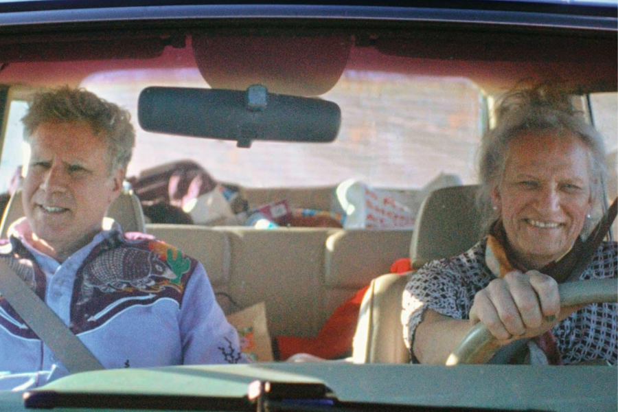 Two people sit in the front seats of a vehicle. The driver smiles while the passenger appears concerned.