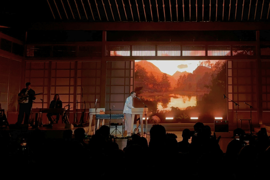 An artist sings into a microphone on a stage made to look like a minka-style house with sliding doors open to reveal a mountain lake scene. A few musicians play in the corner.