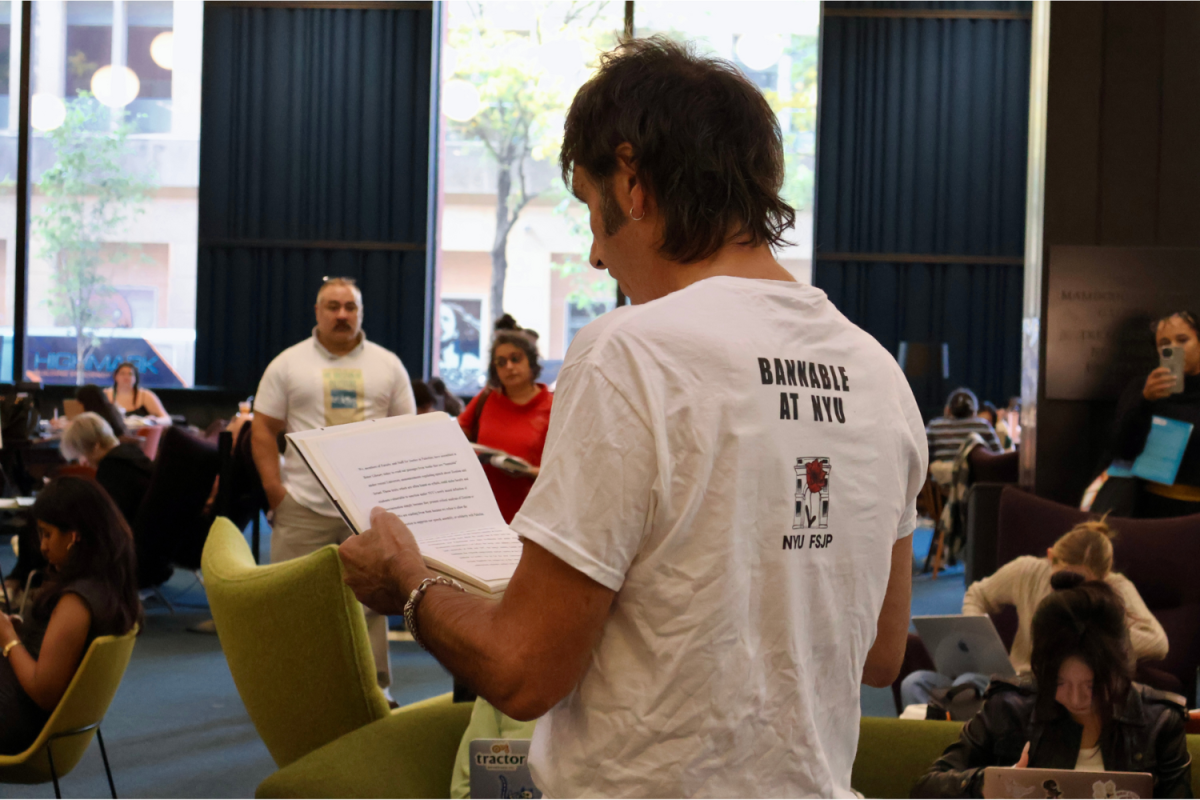 A man reading off a paper in front of people, with “BANNABLE AT N.Y.U.” written on the back of his shirt.