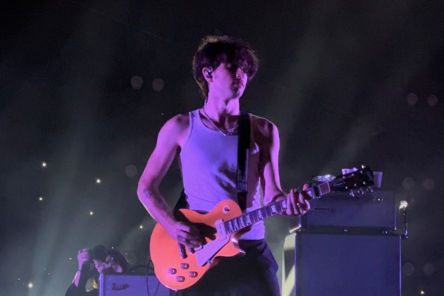 A singer in a white sleeveless shirt performs on stage with an electric guitar under purple and white lights.