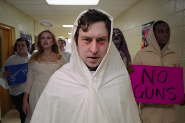 A man wearing a white poncho stands in a hallway. People behind him hold “no guns” signs and are also dressed in white.