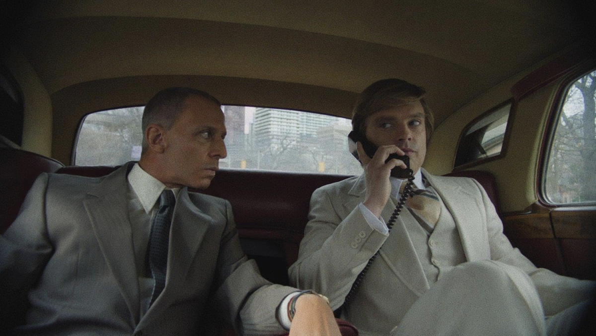 Two men in suits sit in a car. The man on the right is on the phone.