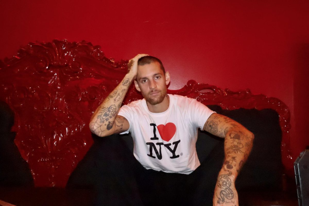 A man wearing a shirt that says “I heart N.Y.” while sitting on a couch in a red room.