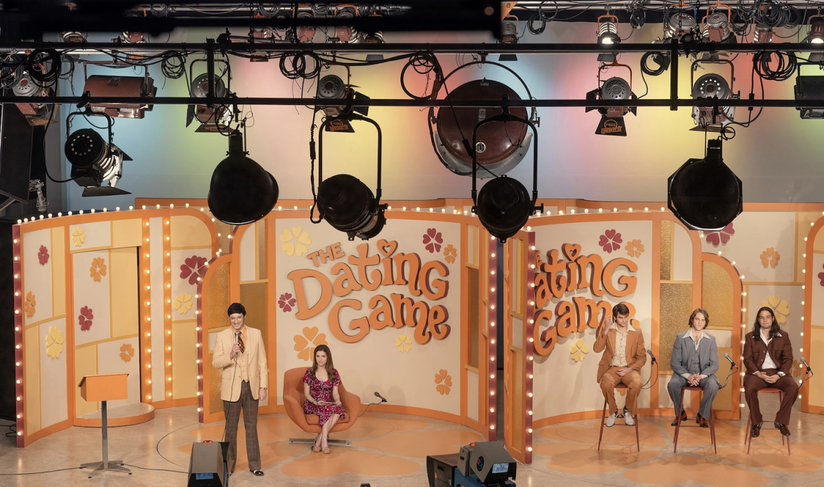 Photographed from behind hanging stage lights is an orange, white and pink stage set with the words “THE DATING GAME.” On one side of a partition is a standing man holding a microphone and a woman in an orange armchair. On the other side are three men sitting on stools.