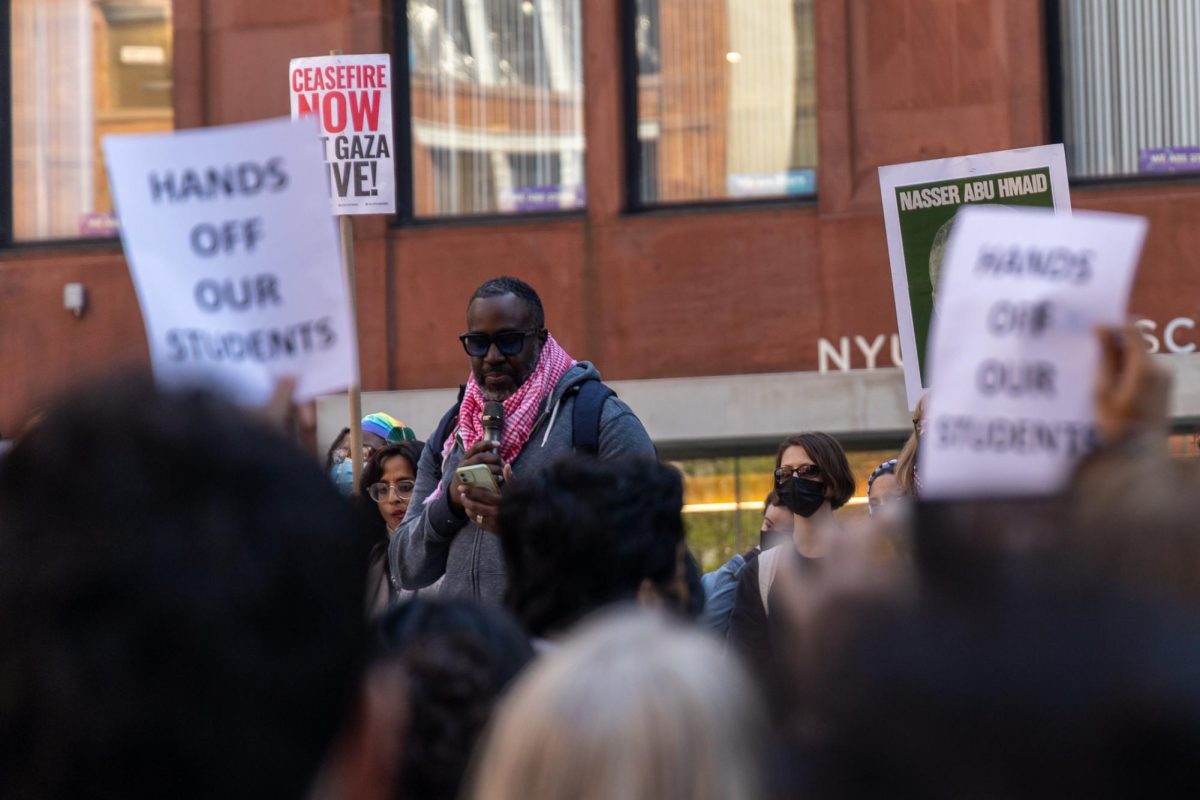 A person outside of N.Y.U. Stern, wearing a red keffiyeh, speaks into a microphone. Around him are protestors holding signs that say “HANDS OFF OUR STUDENTS” and “CEASEFIRE NOW”.