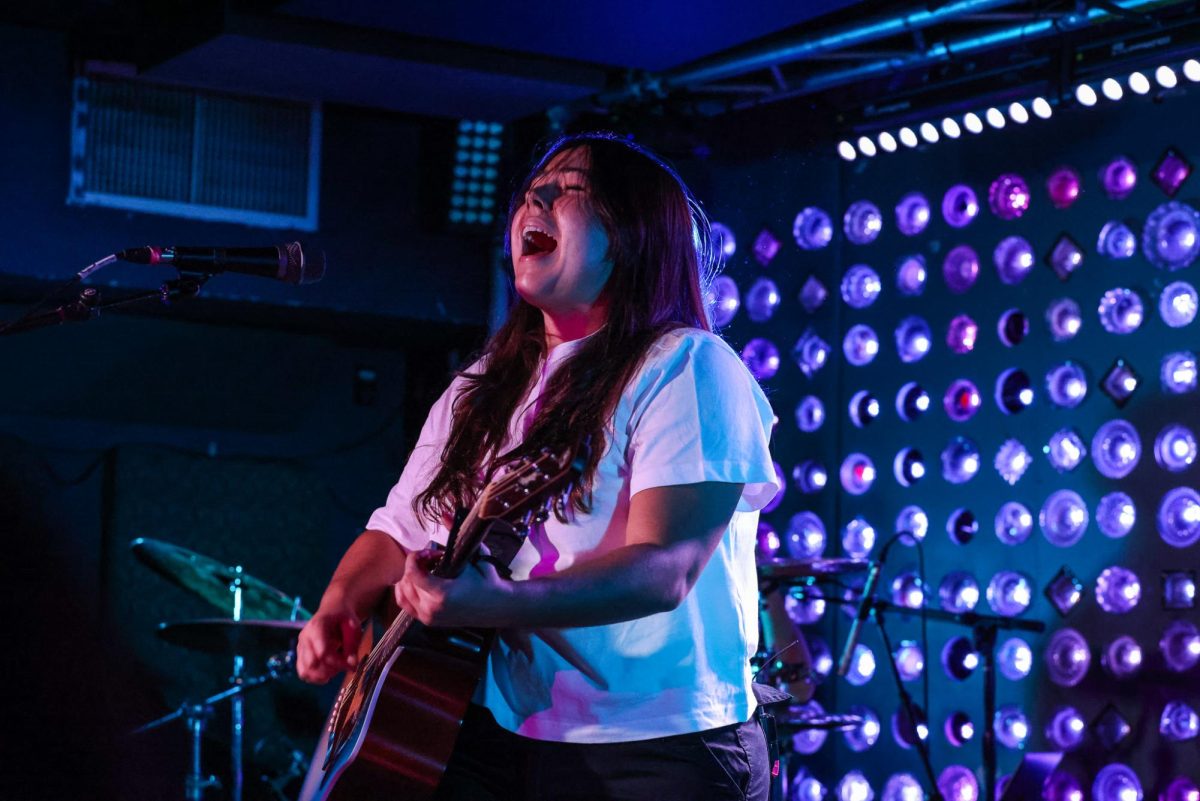 A musician is singing and playing guitar under purple lights during a concert.