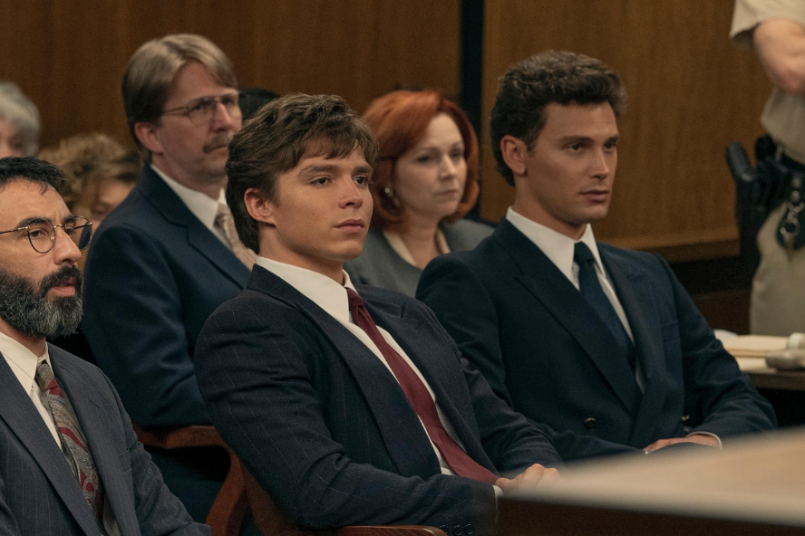 Three men in suits sit at a table in the front of a courtroom. Behind them are more people.