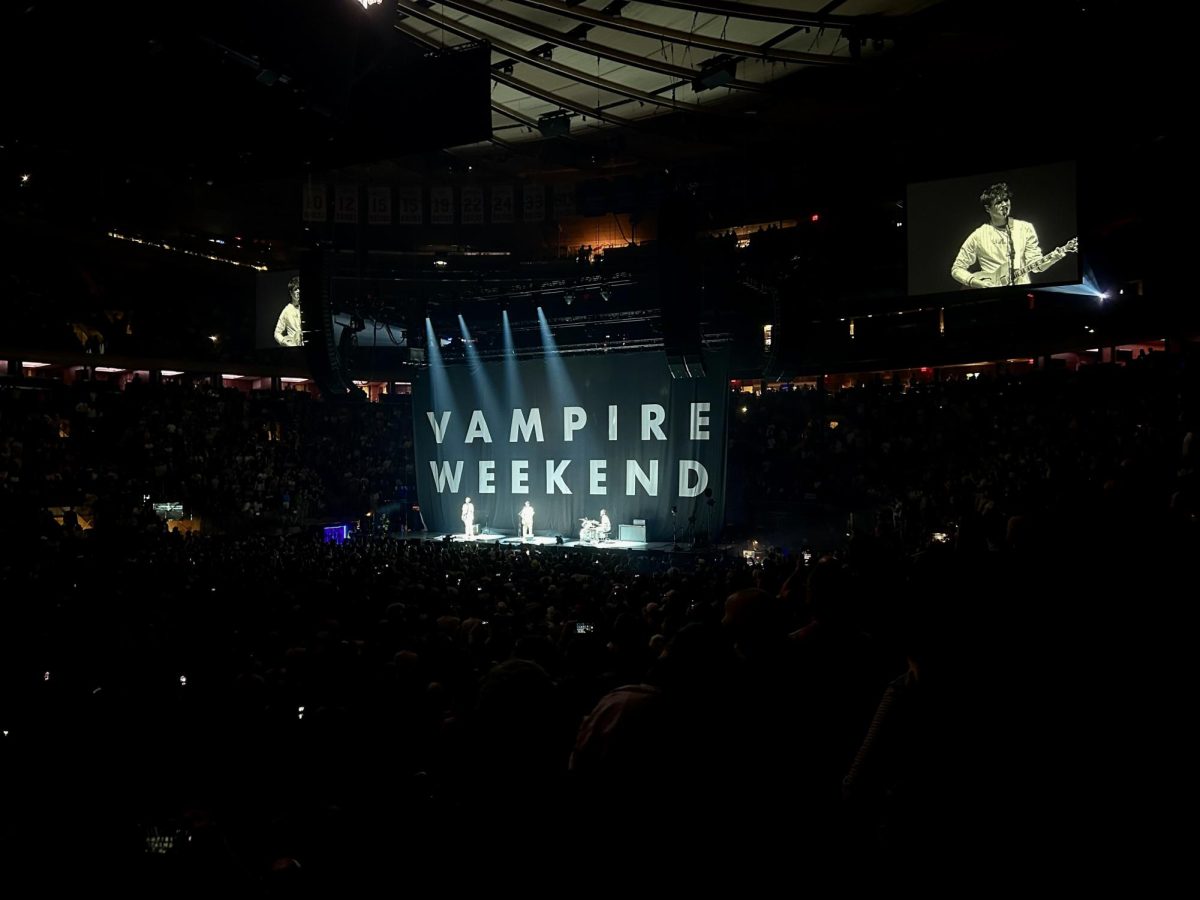 A large crowd in front of a stage with three people in front of a backdrop that says “VAMPIRE WEEKEND." Two screens show close-up shots of a man holding a guitar.