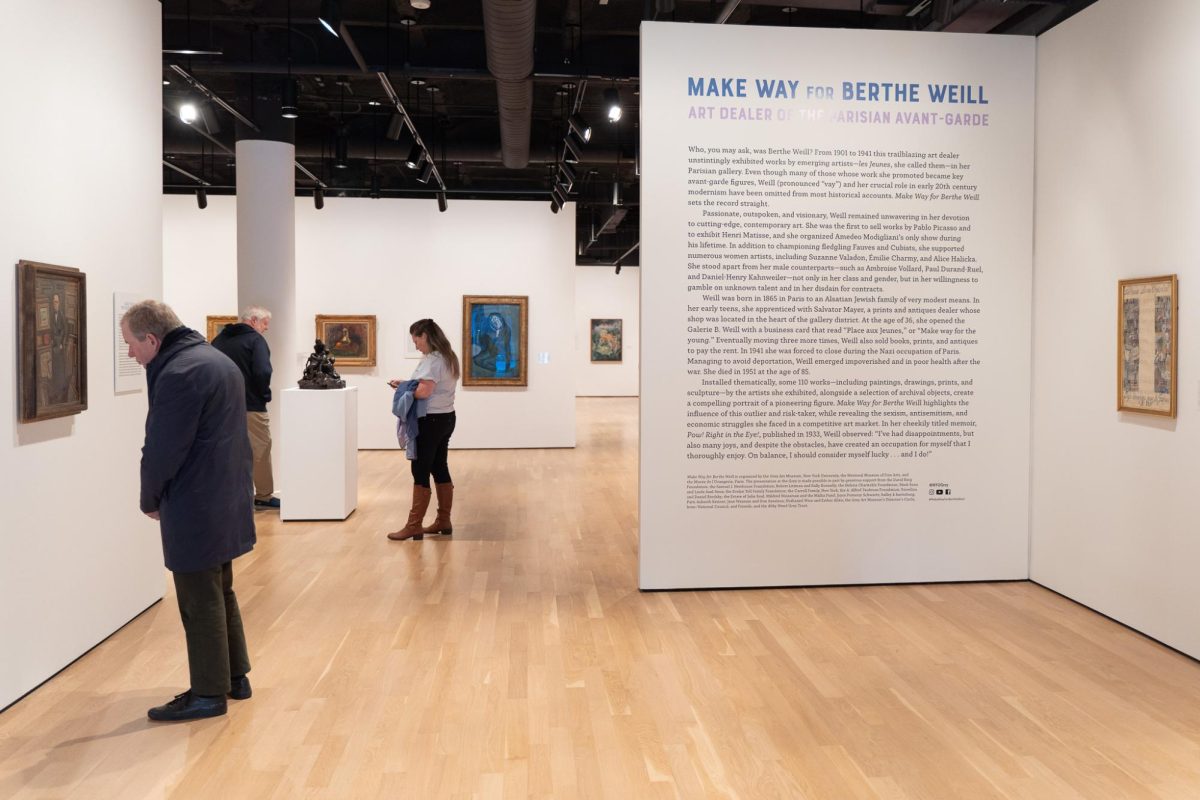 People look at artwork in a gallery. One wall contains a biography of the exhibiting artist titled “Make Way For Berthe Weill” and “Art Dealer Of The Parisian Avant-Garde.”