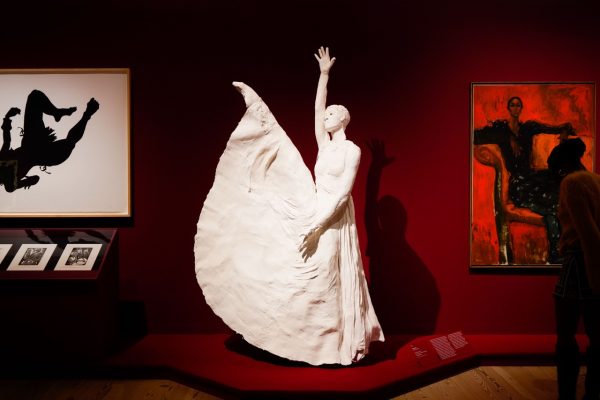 In a dimly lit room, a white sculpture of a dancer with her leg extended in the air and her arm raised up sits on a red platform surrounded by other paintings on the wall. 